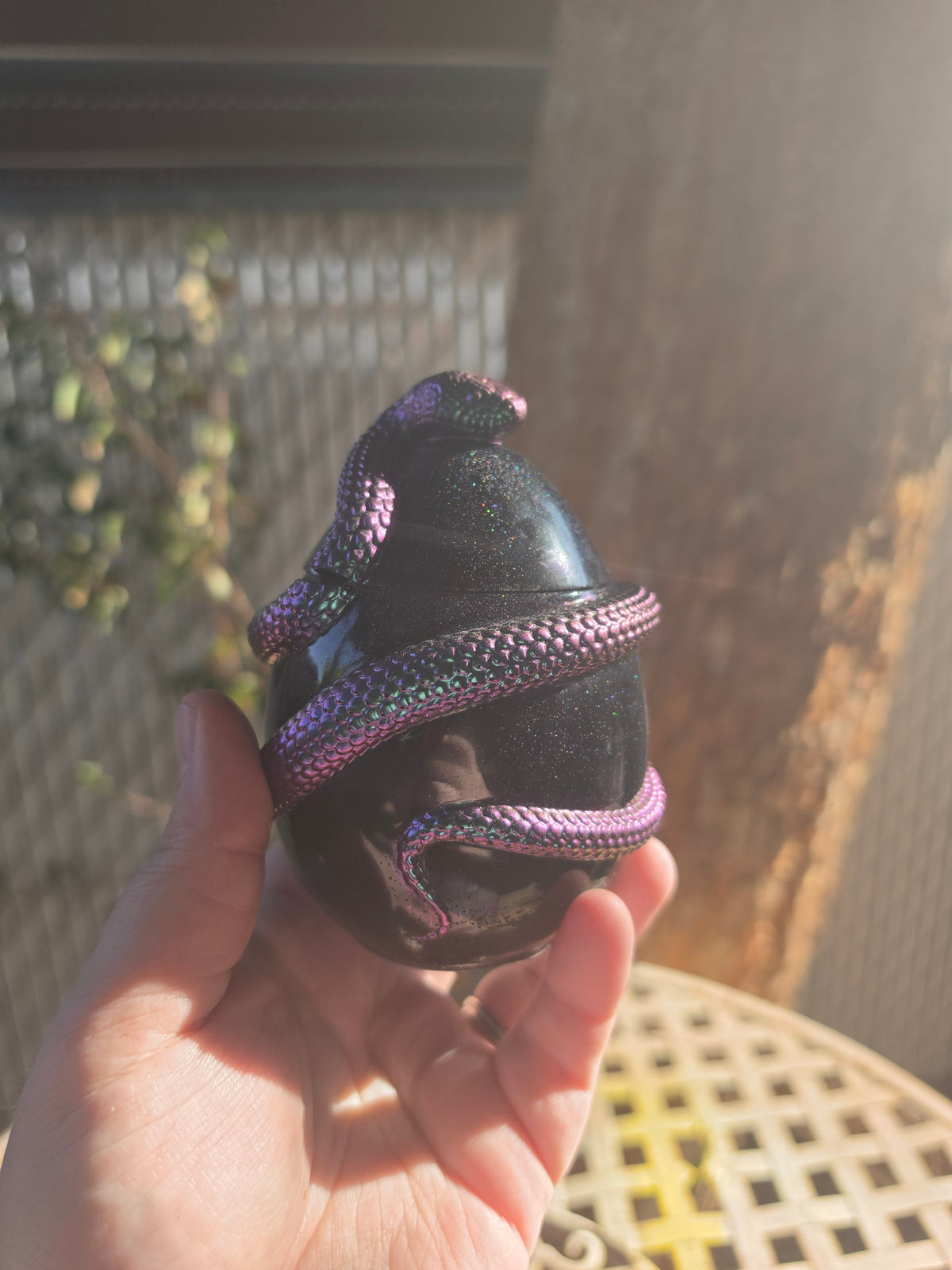 Handmade black resin jar with iridescent purple snake coiled around it, held in hand outdoors
